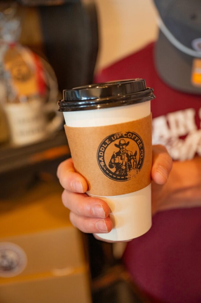 A hand holds a white to-go coffee cup with a black lid and a kraft sleeve stamped with the Saddle Up Coffee Co. cowboy logo