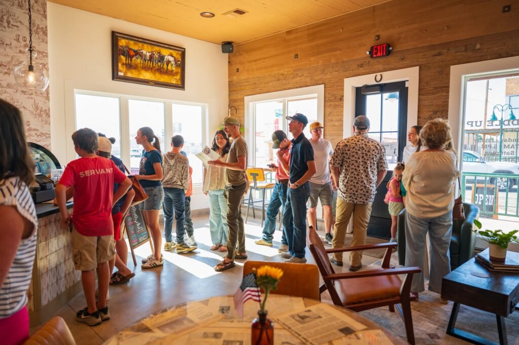 Customers of all ages line up at the order counter inside Saddle Up Coffee Company, a sunny room with wood-plank walls, big windows and a western painting of cattle; in front, a table topped with old newspapers holds a small American flag and a sunflower