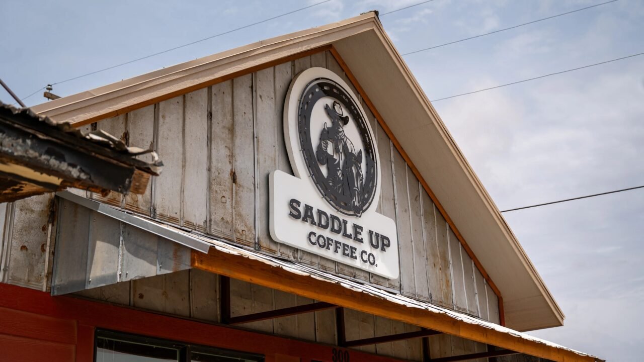 The round black-and-white Saddle Up Coffee Co. sign, with a cowboy holding a coffee cup beside his horse, on the weathered board-and-batten gable of the shop above a rust-orange awning and red walls, under a blue sky