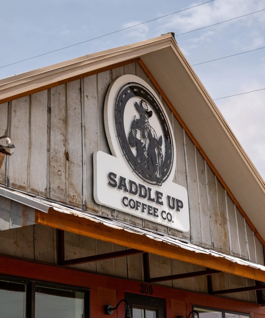 A closer view of the Saddle Up Coffee Co. cowboy logo sign on the weathered wooden gable above the shop's awning and the street number 300