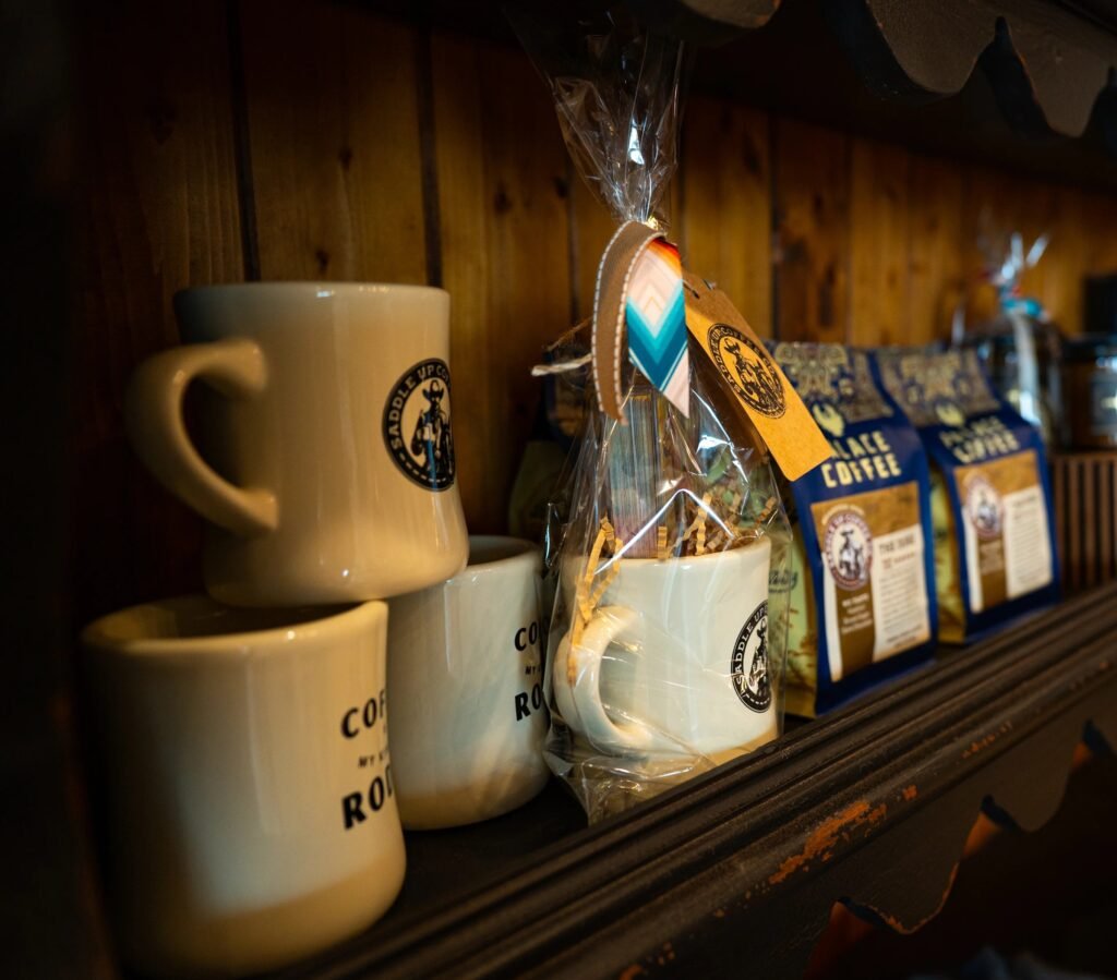 A wooden shelf with cream Saddle Up Coffee Co. logo mugs, a mug gift set wrapped in cellophane with a tag, and blue bags of Palace Coffee beans