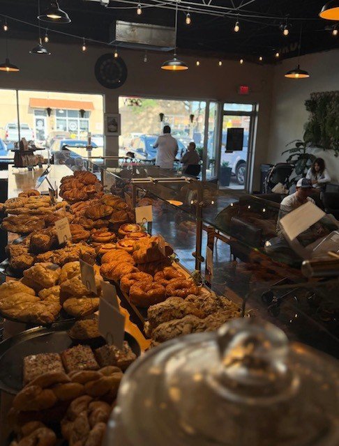 A bakery display case filled with croissants, danishes, sticky buns and scones, with customers in the background of a bright cafe lit by pendant lights.