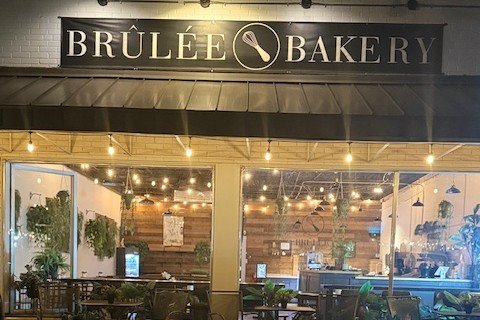 Brûlée Bakery storefront at night with a black sign bearing the bakery name and a whisk logo, string lights, patio tables and chairs, and potted plants in front of the glass windows.