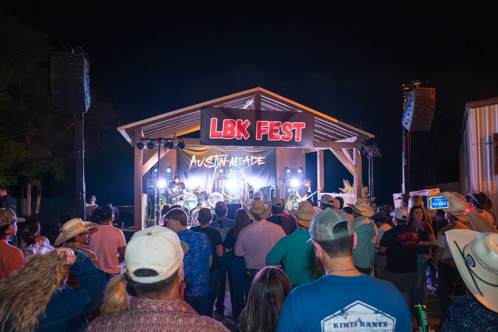 A crowd in cowboy hats and caps faces a lit pavilion stage with a red-and-white LBK FEST banner and an Austin Meade backdrop at night.