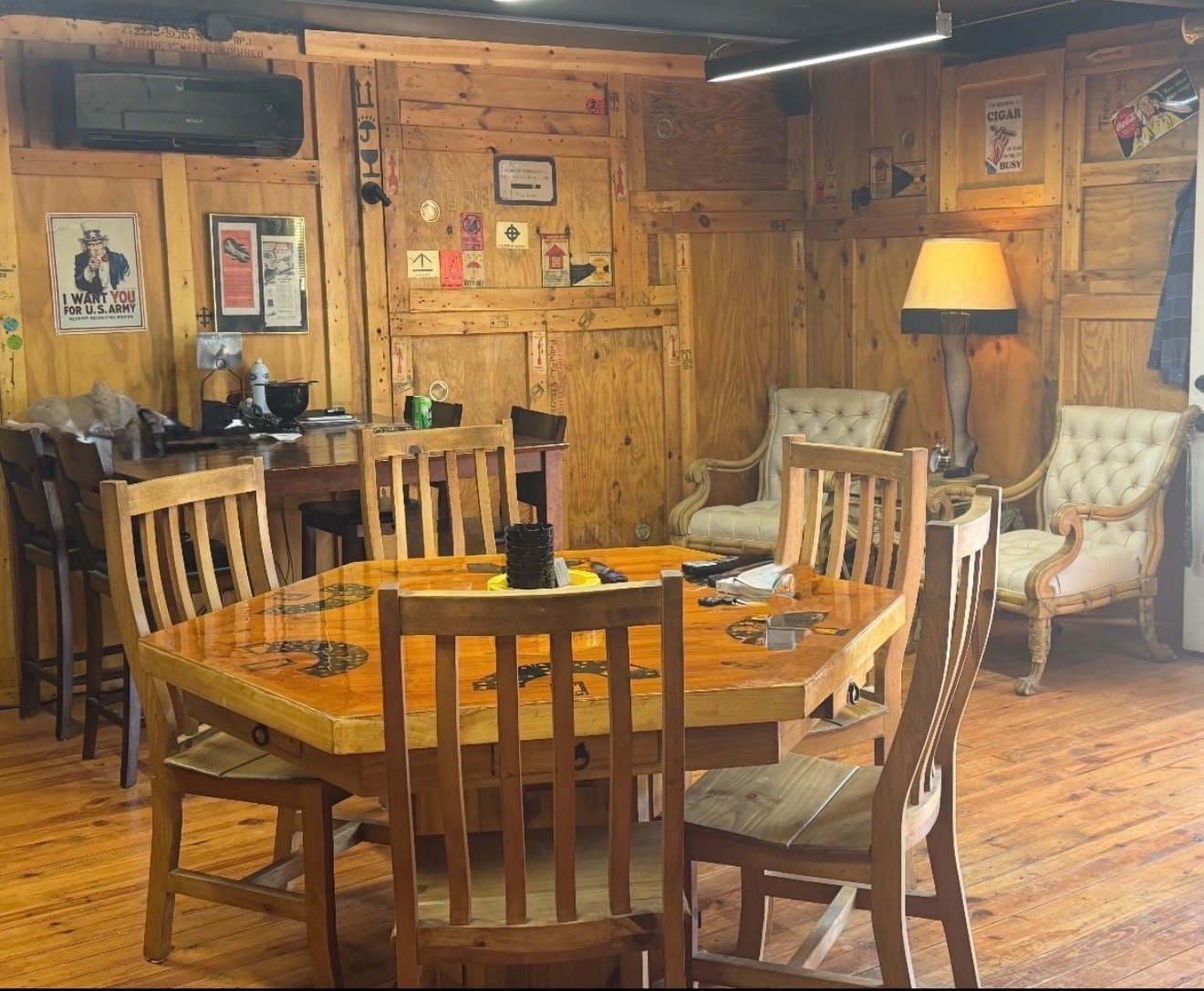 The lounge at Mystery Shack Cigars & Coffee, with wood-paneled walls, a large wooden game table with chairs, two tufted cream armchairs, a leg-lamp floor lamp and vintage posters.