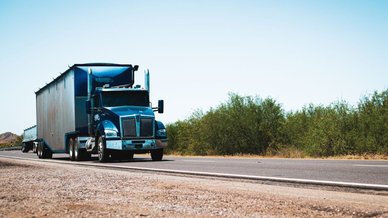 A blue semi-truck on an open highway under a clear blue sky.