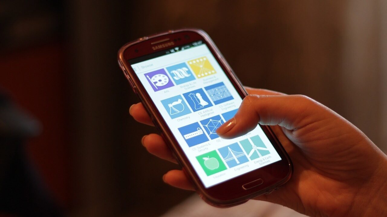 A person's hand holding a smartphone showing a screen of app icons, against a dark blurred background