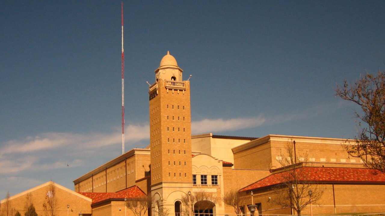 The tan brick bell tower and entrance of United Supermarkets Arena at Texas Tech University in Lubbock under a blue sky