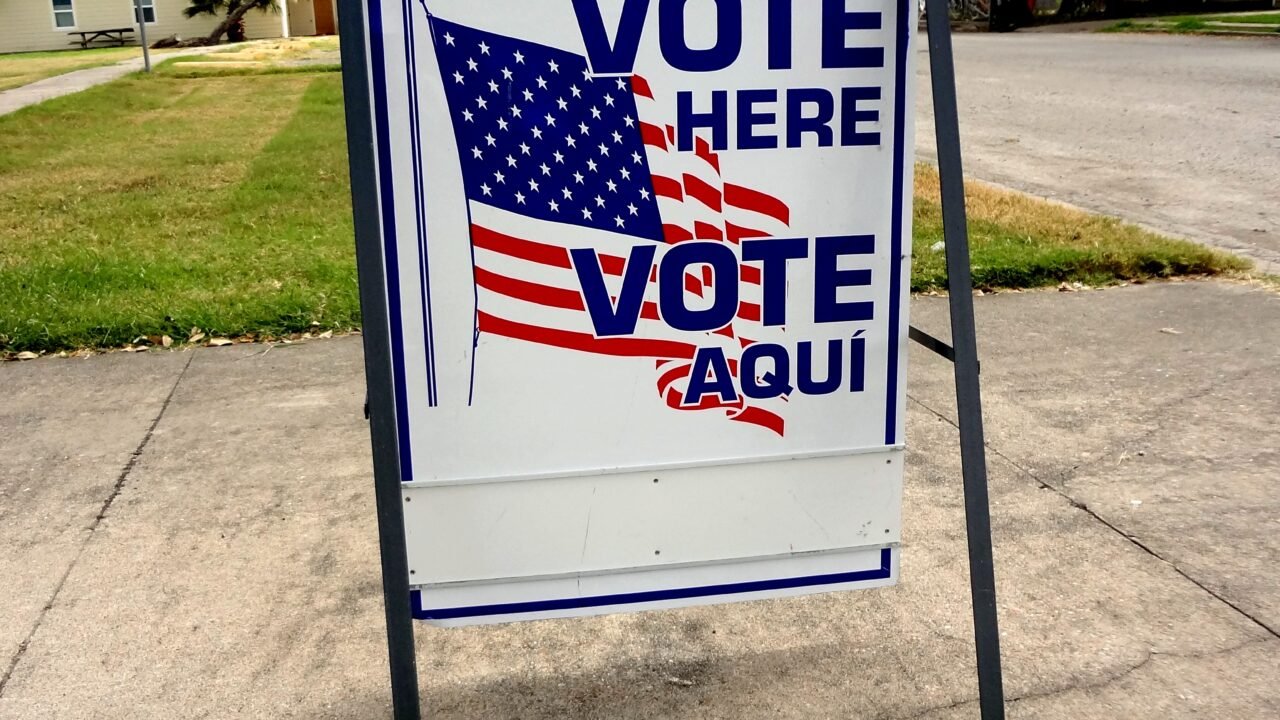A bilingual "Vote Here / Vote Aquí" sign with an American flag stands on a sidewalk outside a polling place.